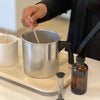 Woman stirring melted wax in a silver pour pot. Amber fragrance bottle and hourglass in foreground. White concrete vessel next to pour pot.