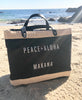 a black jute market bag with Peace + Aloha Makana on the front sitting on the sand at the beach